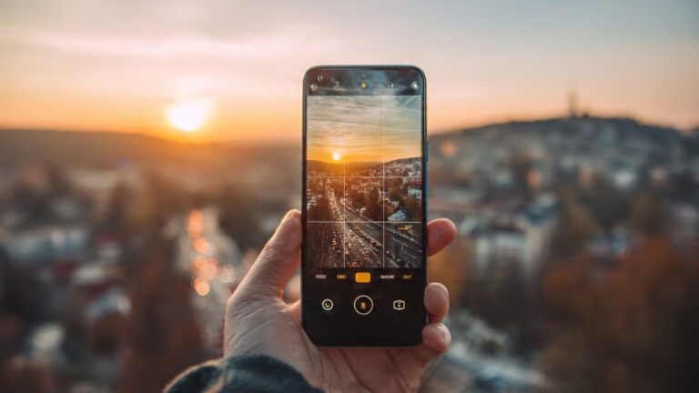 Person holding smartphone taking photo during golden hour with cityscape in background